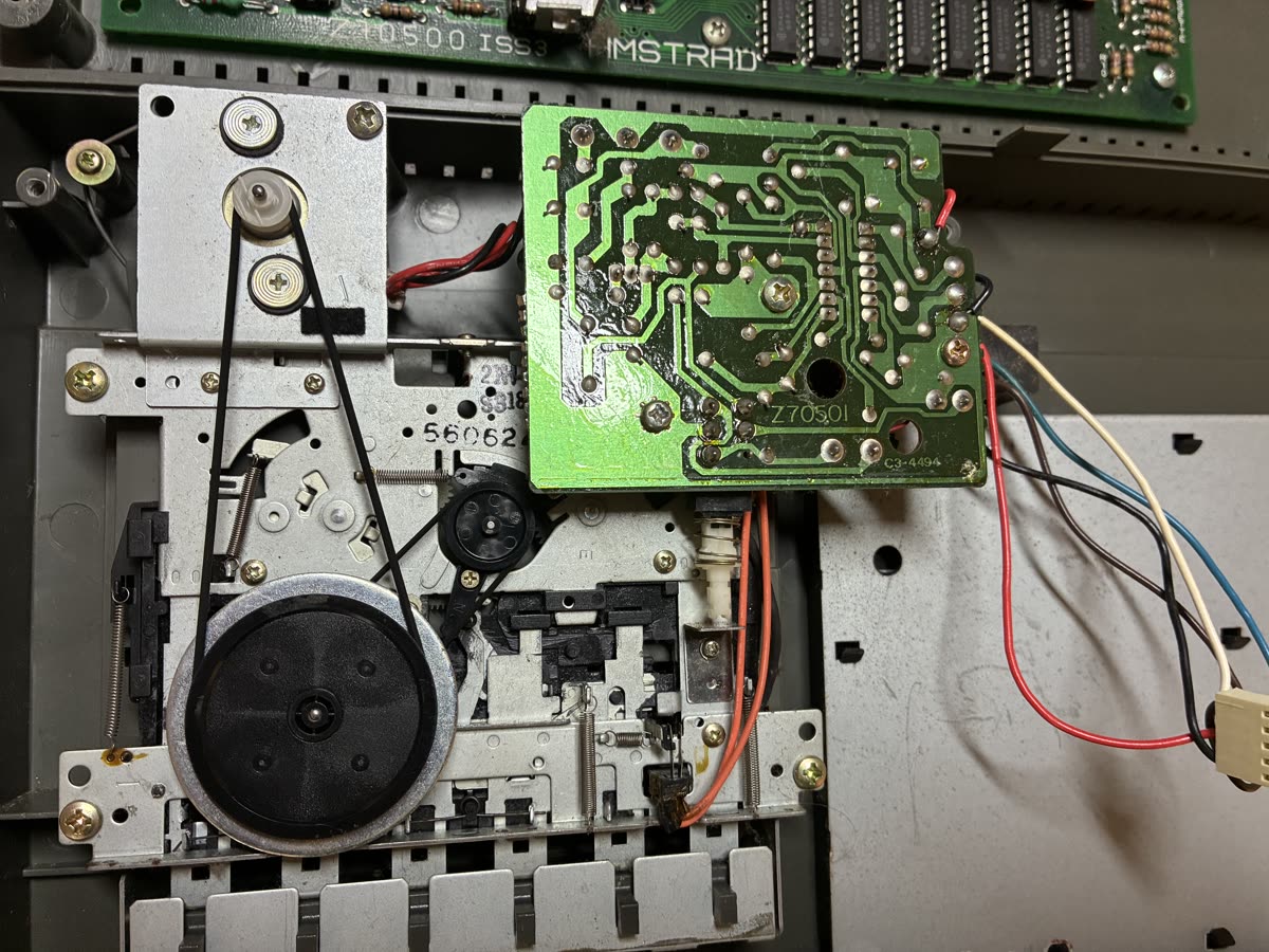 Close-up of the Datacorder mechanism: a large black flywheel with a rubber belt running up to the motor pulley, next to the deck's circuit board.