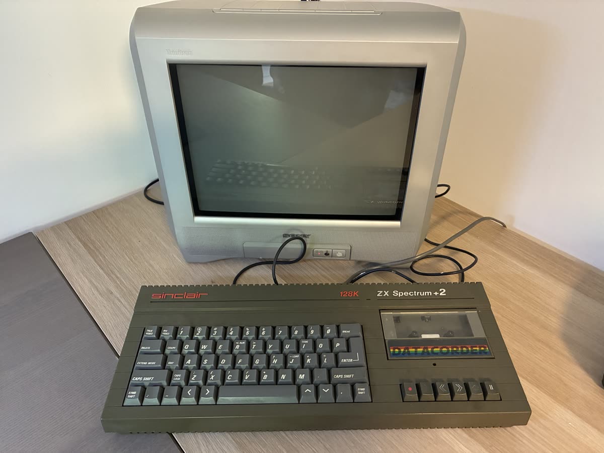 A grey Sinclair ZX Spectrum +2 128K with its built-in Datacorder cassette deck, in front of a small silver Sony CRT television.